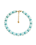 A beaded necklace displayed on a cream background with soft leaf shadows. The necklace features light aqua blue round beads alternating with smaller turquoise blue disc beads, finished with a gold clasp and extender chain.