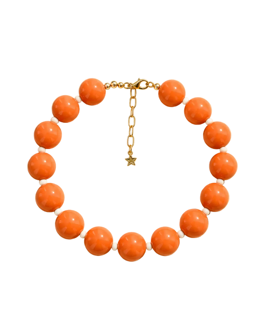 A beaded necklace with large round orange-brown beads separated by small white beads, with a gold chain clasp and star charm, displayed on a beige background with leaf shadows. Text above and below reads 'STACKED BY VALE' and 'CAPRI POP NECKLACE'.